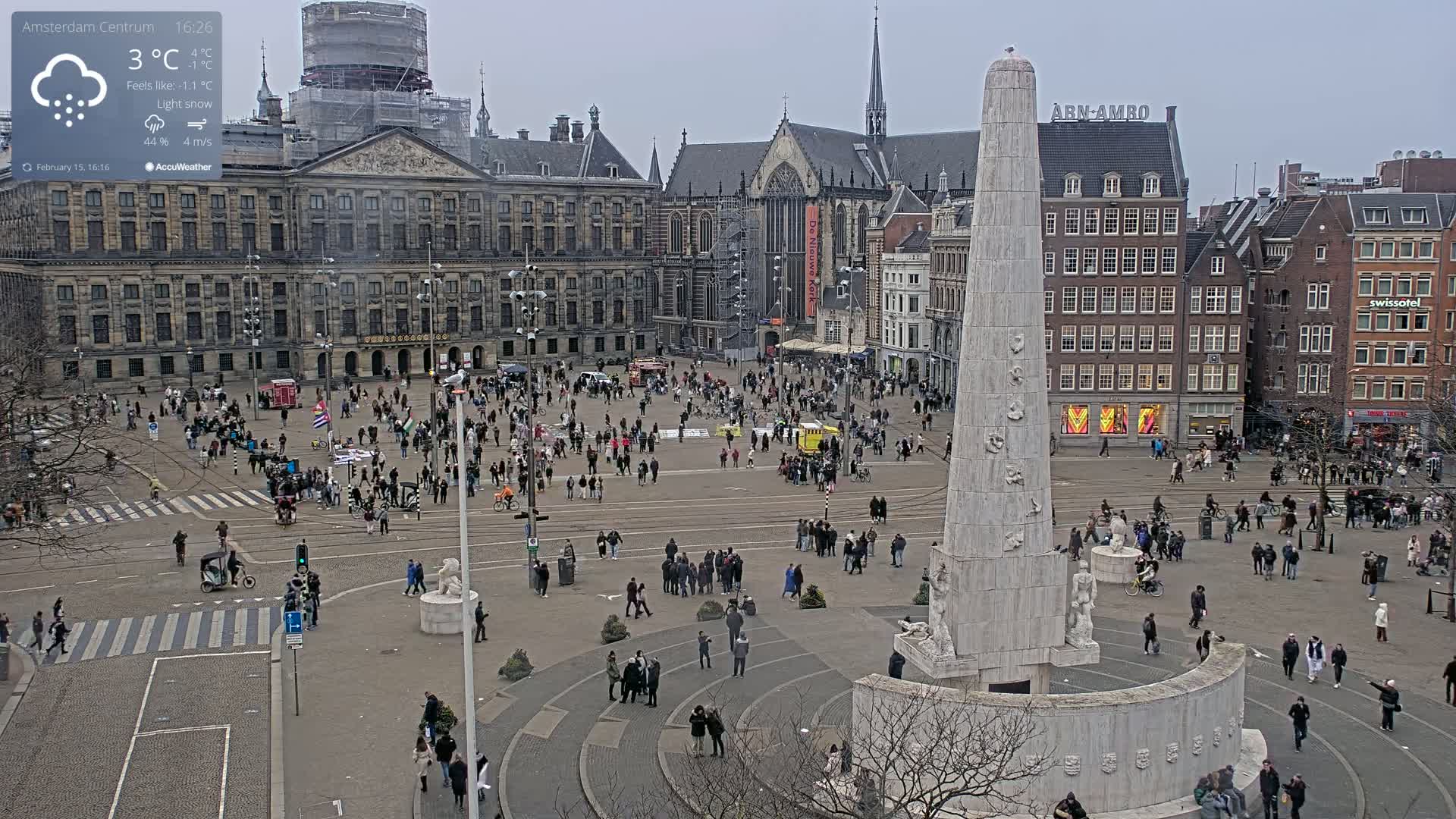 A large, crowded city square, under a light snow, features a tall monument at its center and several historic buildings surrounding it.
