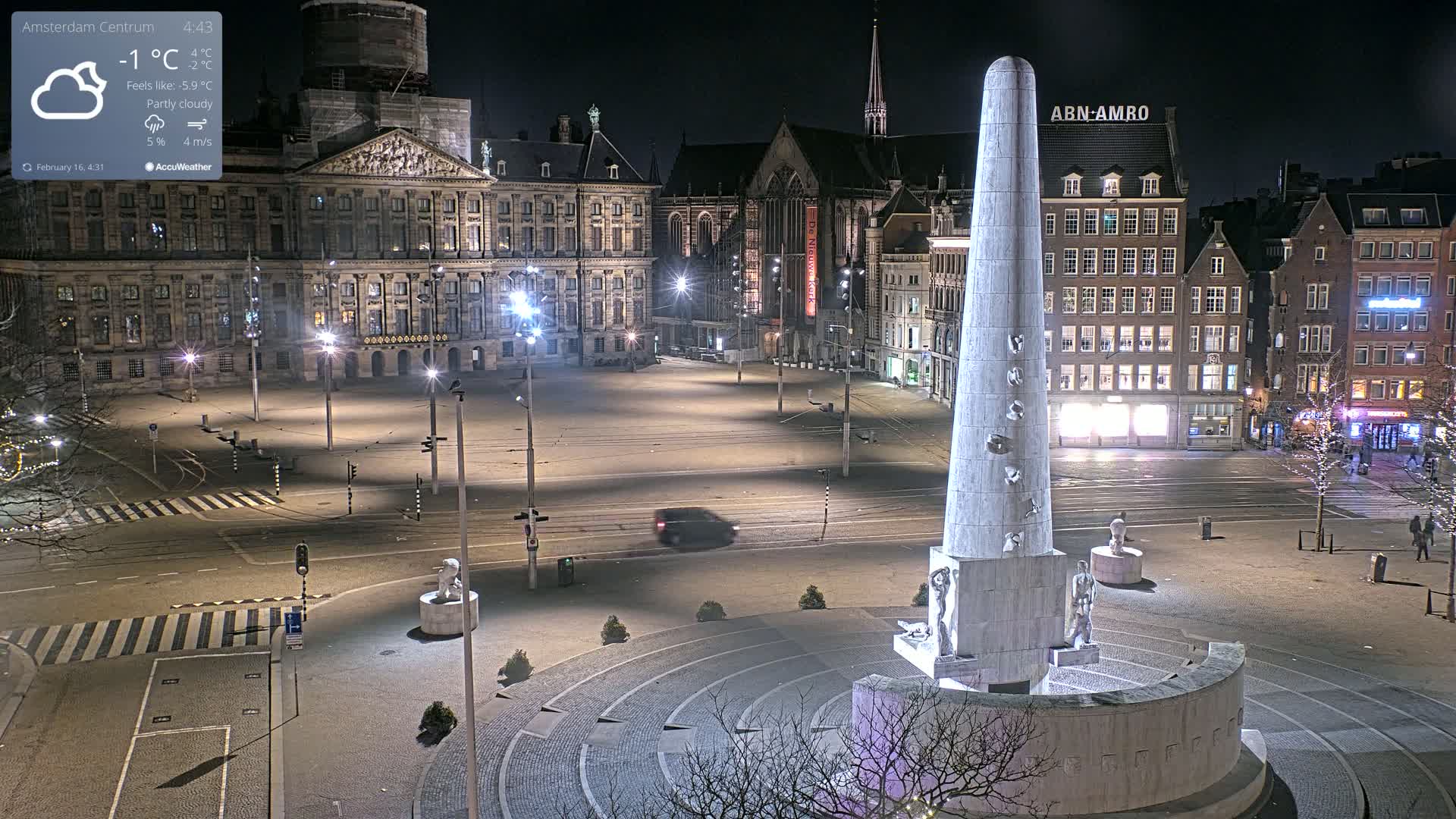 A mostly empty city square at night under partly cloudy skies, featuring a tall monument at the center and buildings illuminated along the edges.