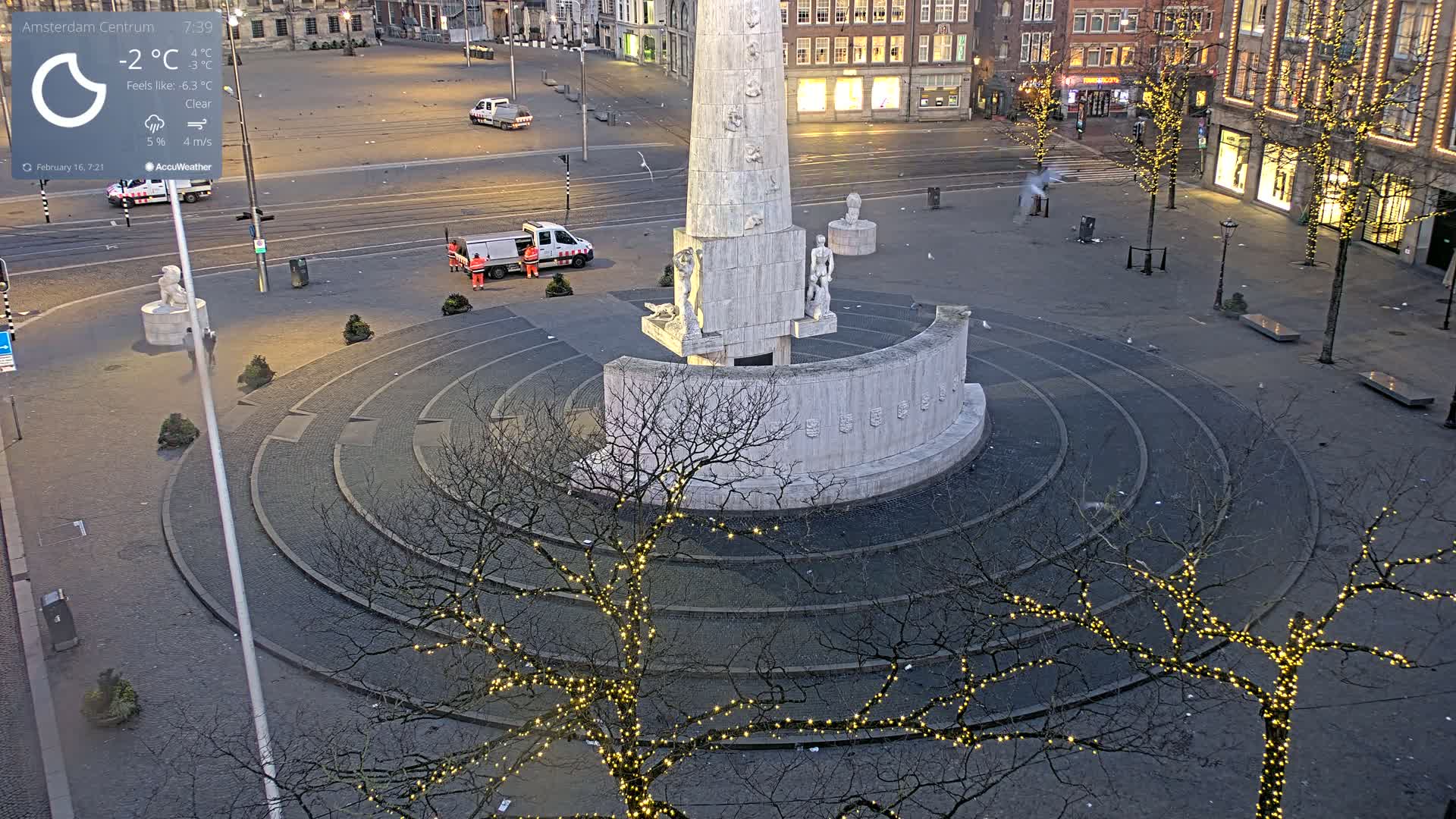 A monument with circular steps and string lights on surrounding bare trees sits in a mostly empty city square under a clear sky at -2°C.