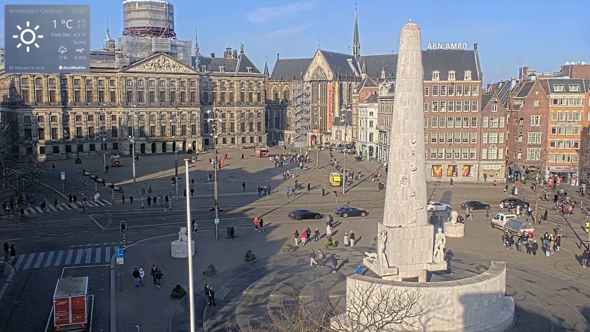 A sunny, but cold, city square with a large monument at the center, surrounded by historical buildings and people walking.