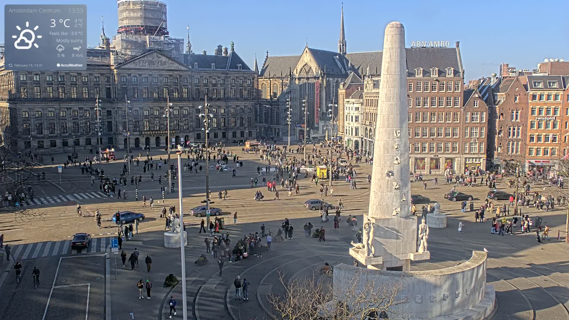 A mostly sunny city square with a tall monument in the center, surrounded by buildings and many people.