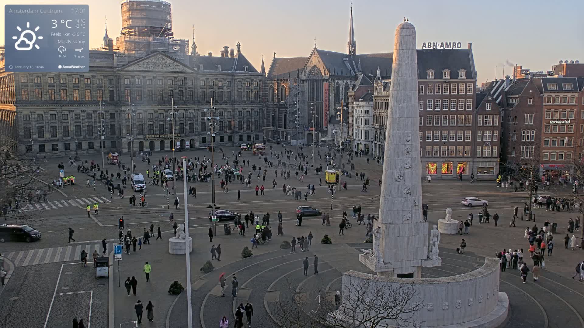 A mostly sunny, cold day in a large European city square, showing many people walking around a tall monument, with various buildings surrounding the open space.