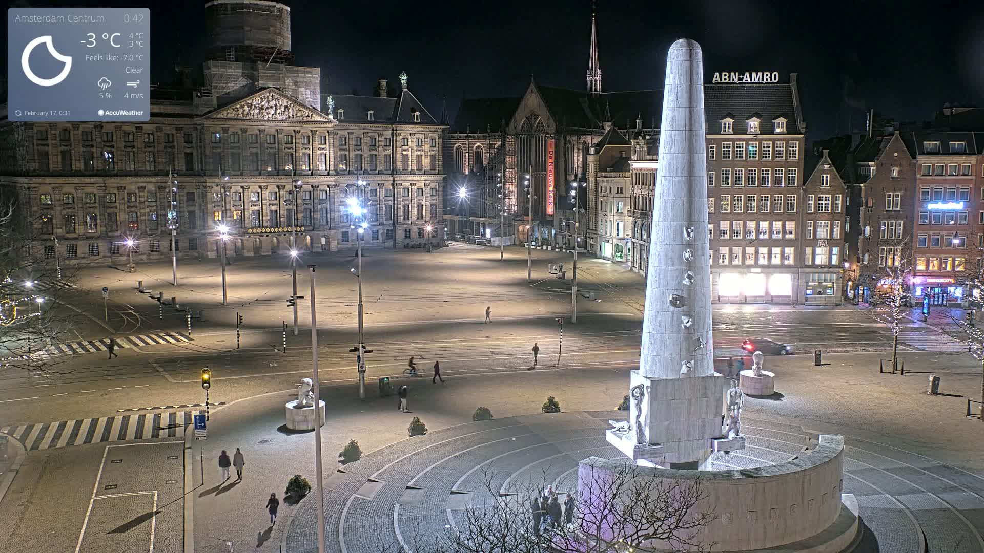 A nighttime, clear view of a large, mostly empty city square with a tall monument at its center, surrounded by buildings and streetlights.