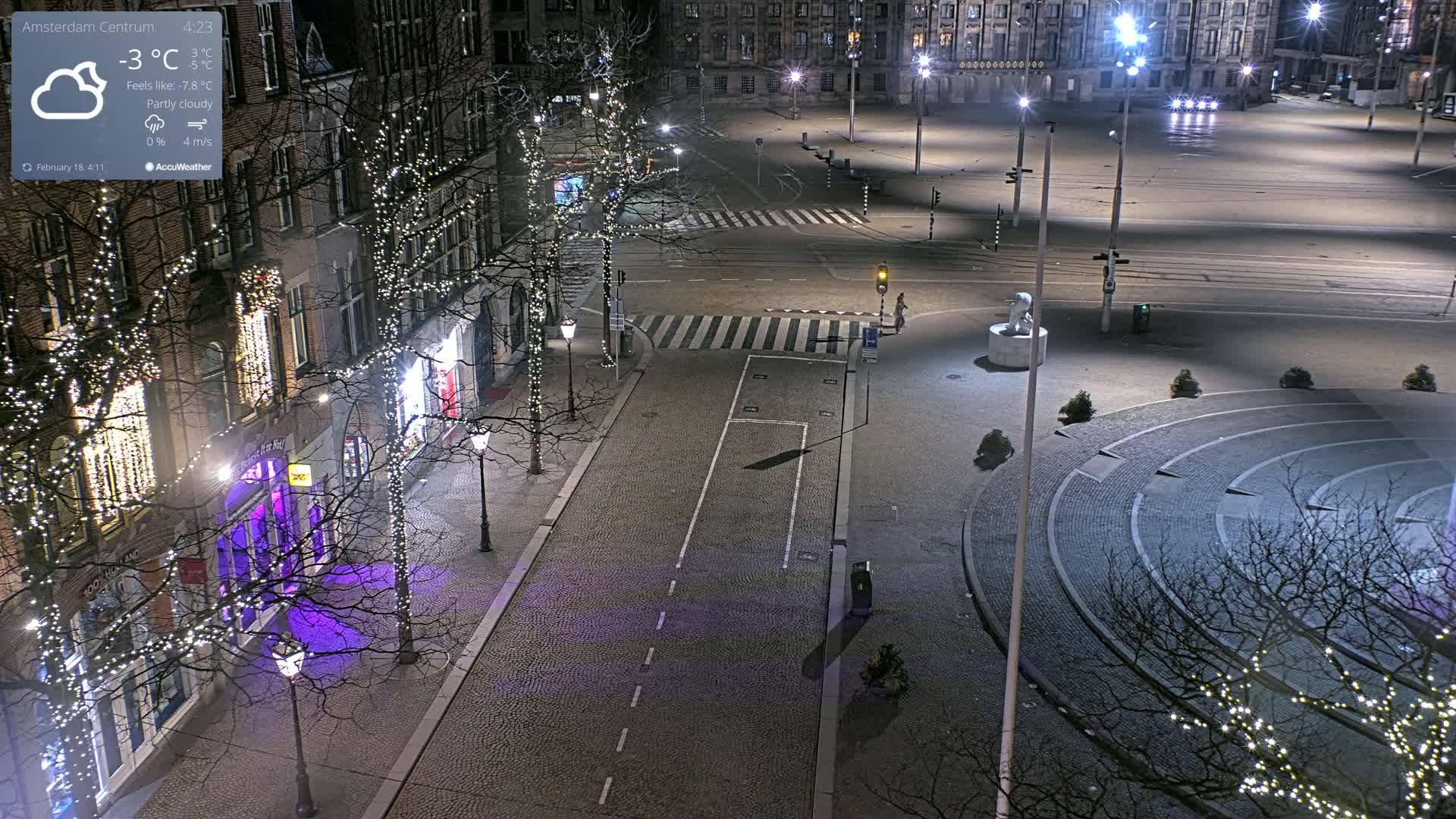 A mostly empty, cobblestone city square at night under partly cloudy skies, illuminated by streetlights and string lights on trees lining the streets.