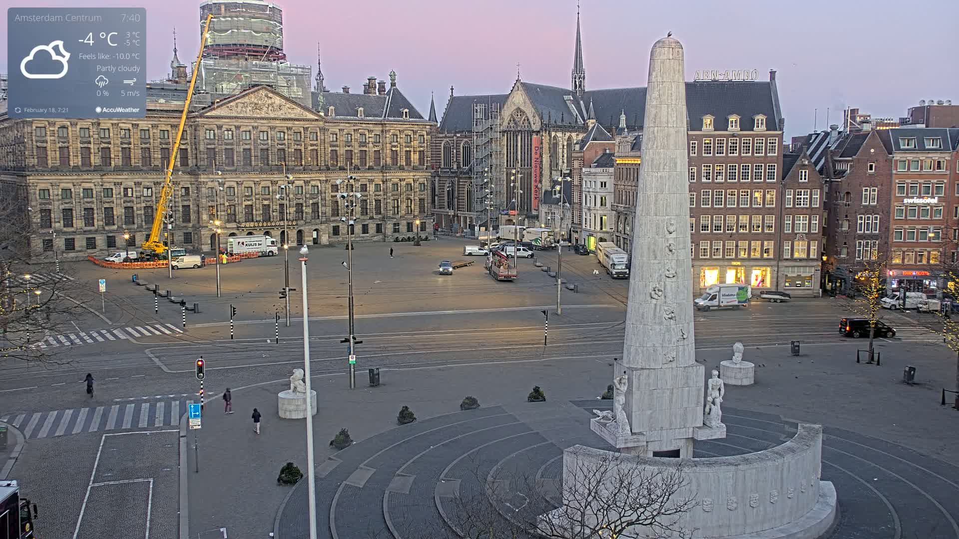 A mostly empty city square at dawn under partly cloudy skies, featuring a large monument in the foreground and several buildings, including a palace and church, in the background.