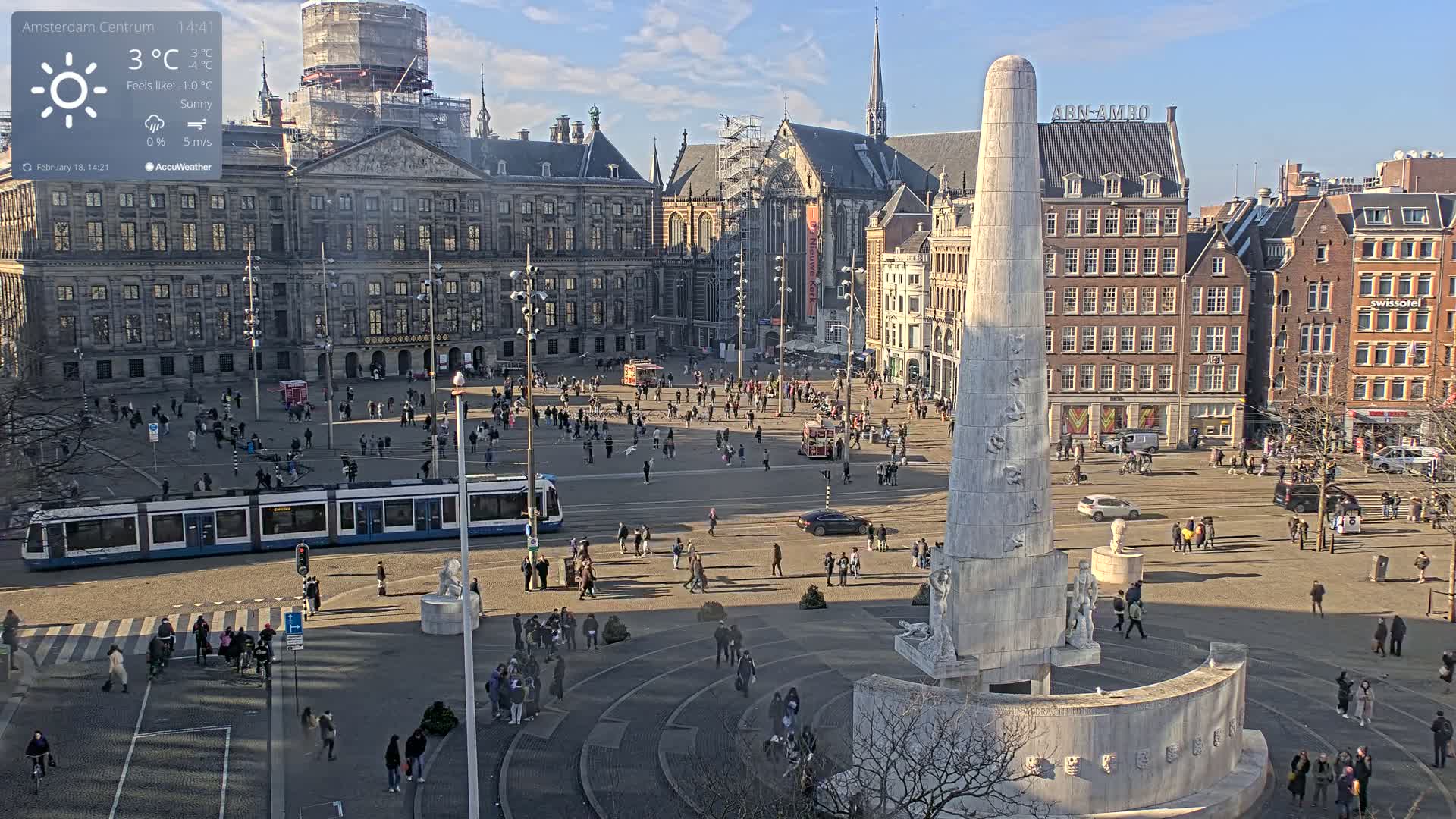 A sunny city square with a tall monument at its center, surrounded by buildings, a tram, and many pedestrians.