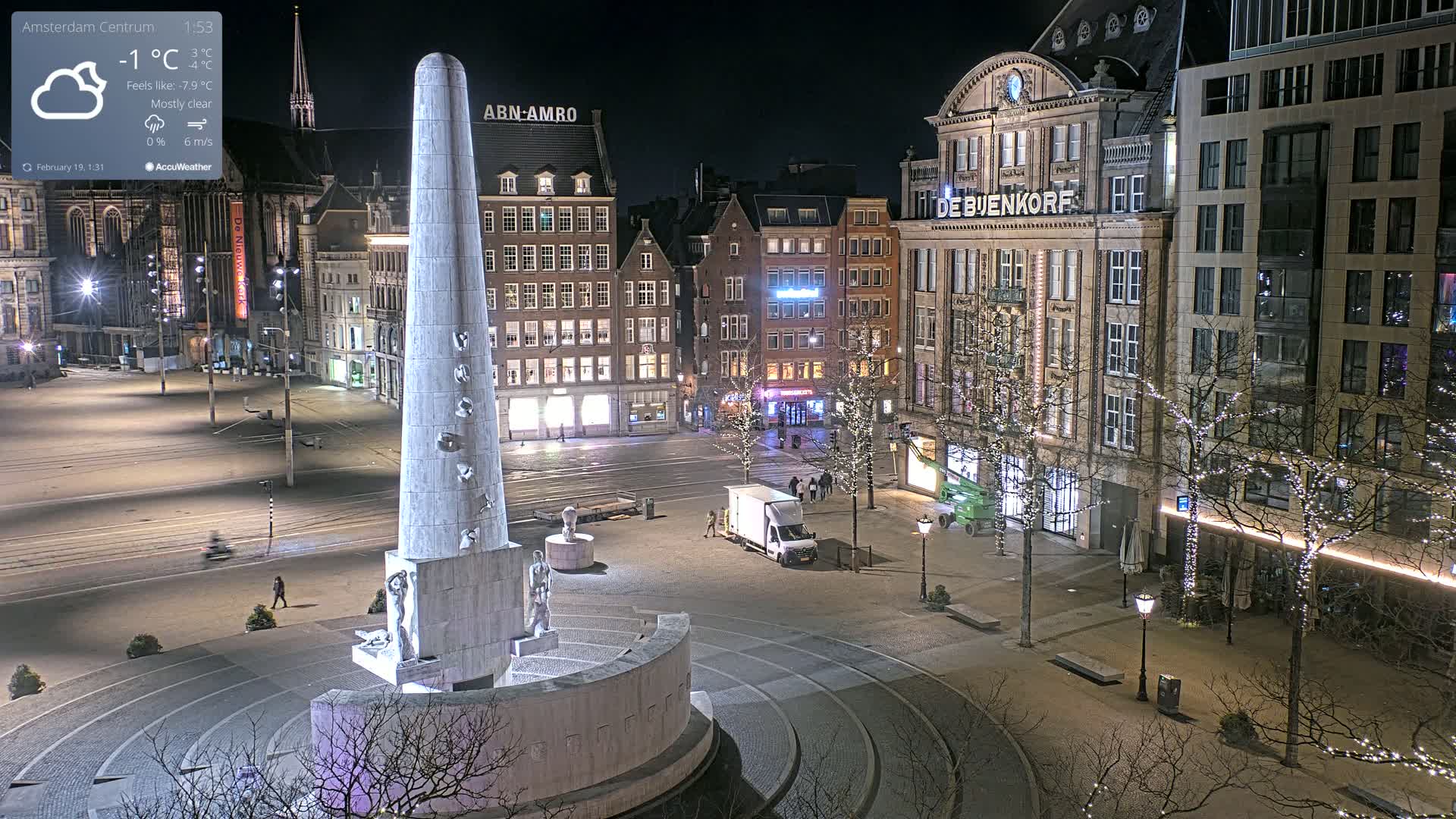 A nighttime view of a city square with a tall, light-colored monument in the foreground, surrounded by buildings decorated with Christmas lights, under mostly clear skies and a temperature of -1°C.
