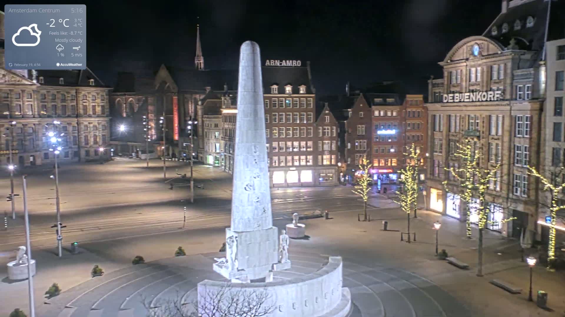 A mostly cloudy night scene shows a tall, slender monument in a city square surrounded by illuminated buildings.