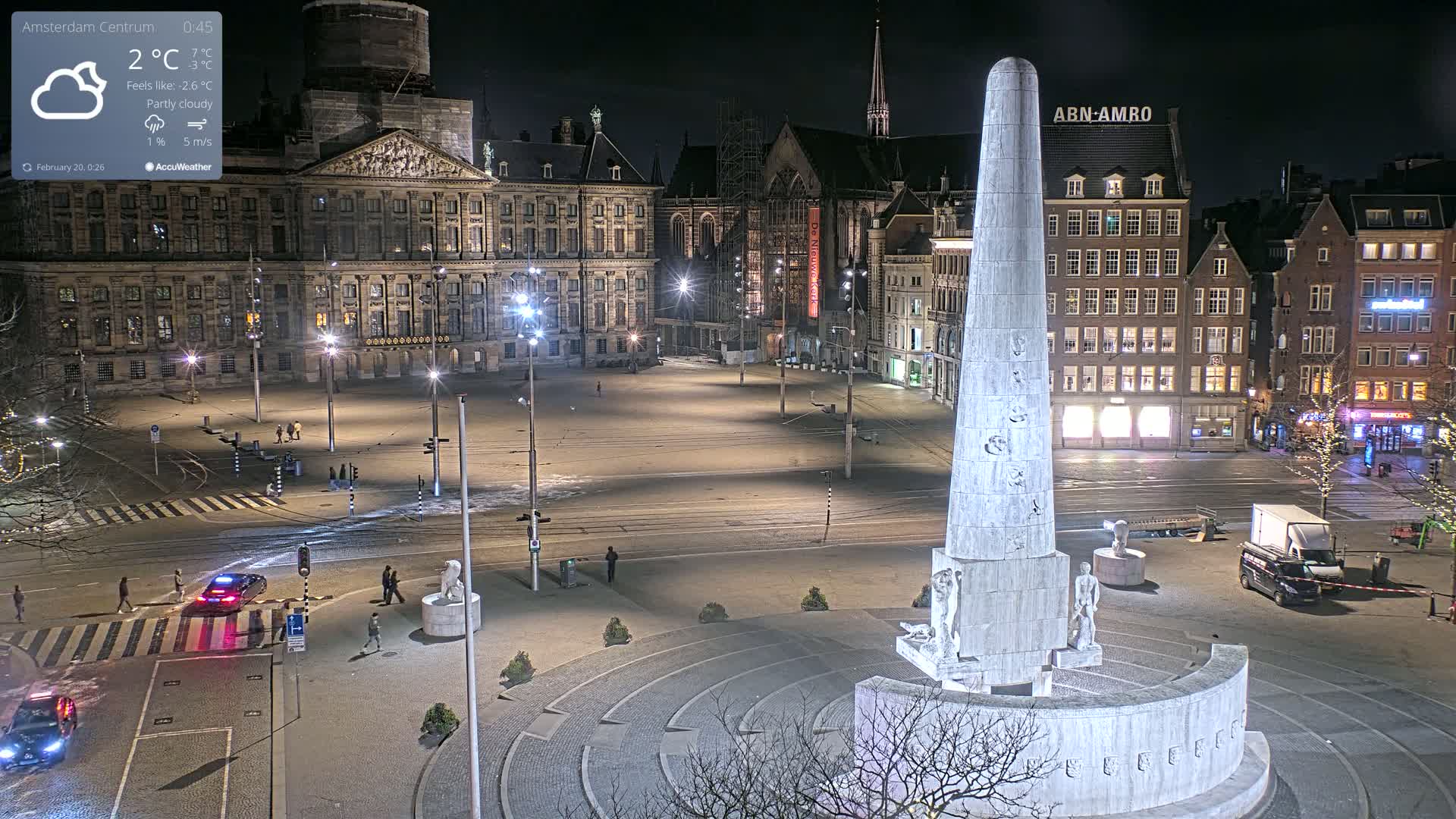 A nighttime, partly cloudy view of a large, mostly empty city square with a tall monument at its center, surrounded by historical buildings and illuminated by streetlights.