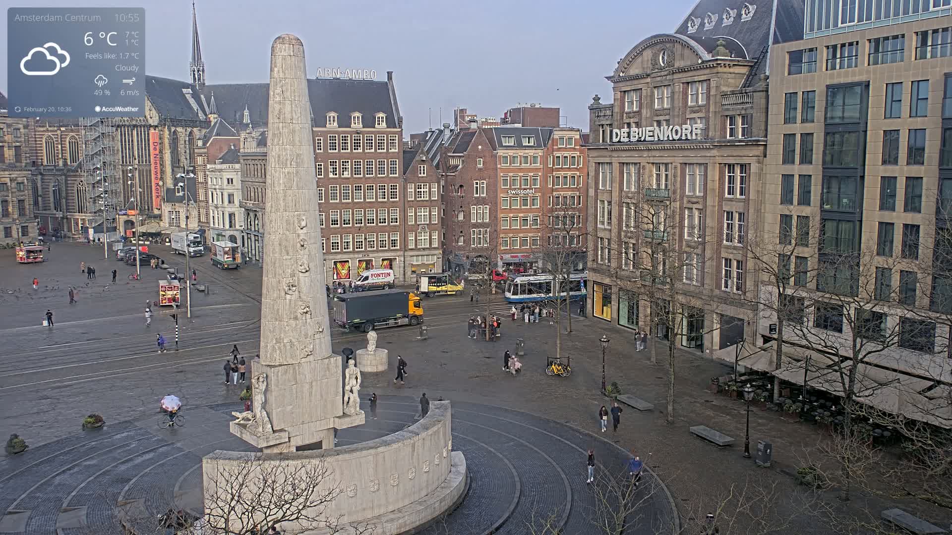 A cloudy day in a city square, centered on a tall stone monument, with people walking among several buildings and a tram.