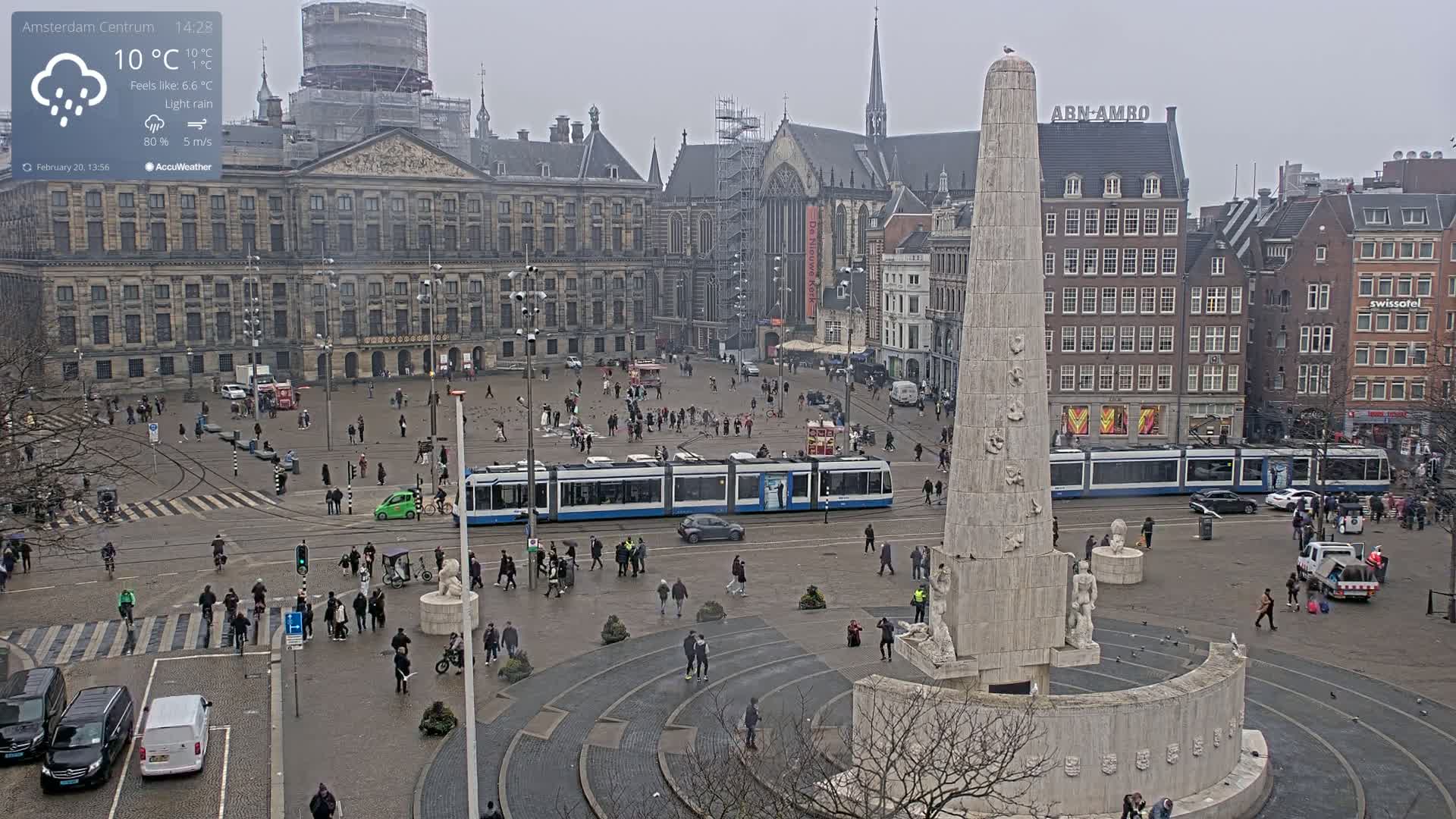 A city square with a tall monument in the center, surrounded by buildings and people, under light rain.