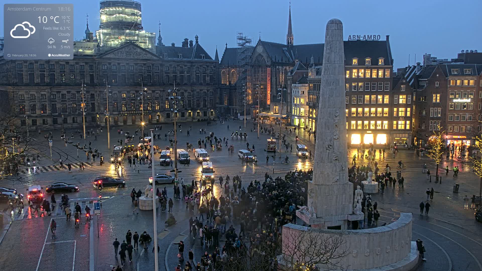 A large city square at dusk under cloudy skies with a light rain shows many people gathered around a tall monument, with cars and buildings surrounding the area.