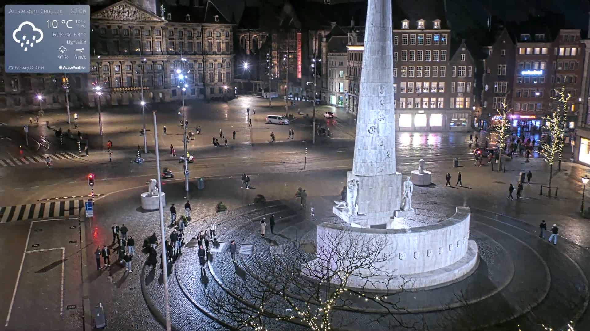 A nighttime view of a city square with a tall monument at its center, under light rain.