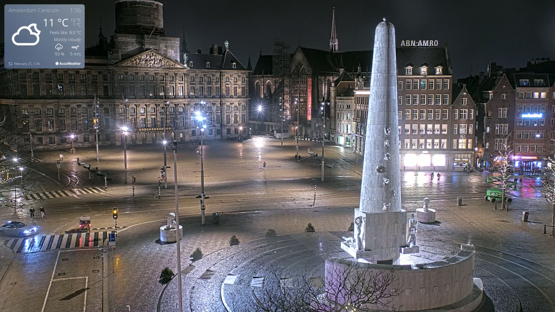 A mostly cloudy night scene shows a large, empty town square with a tall monument in the center, surrounded by buildings and streetlights.