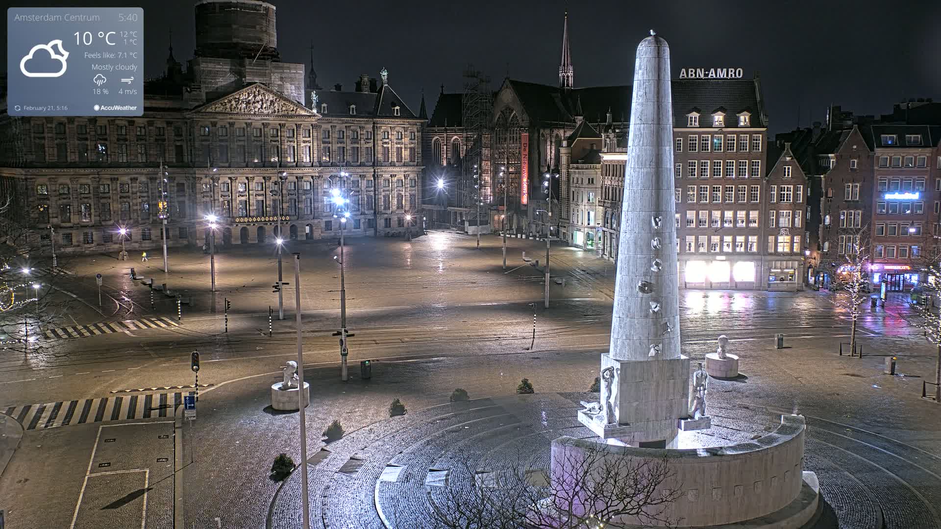 A mostly cloudy night scene shows a large, empty city square with a tall monument at its center, surrounded by historic buildings and wet pavement under illuminated streetlights.