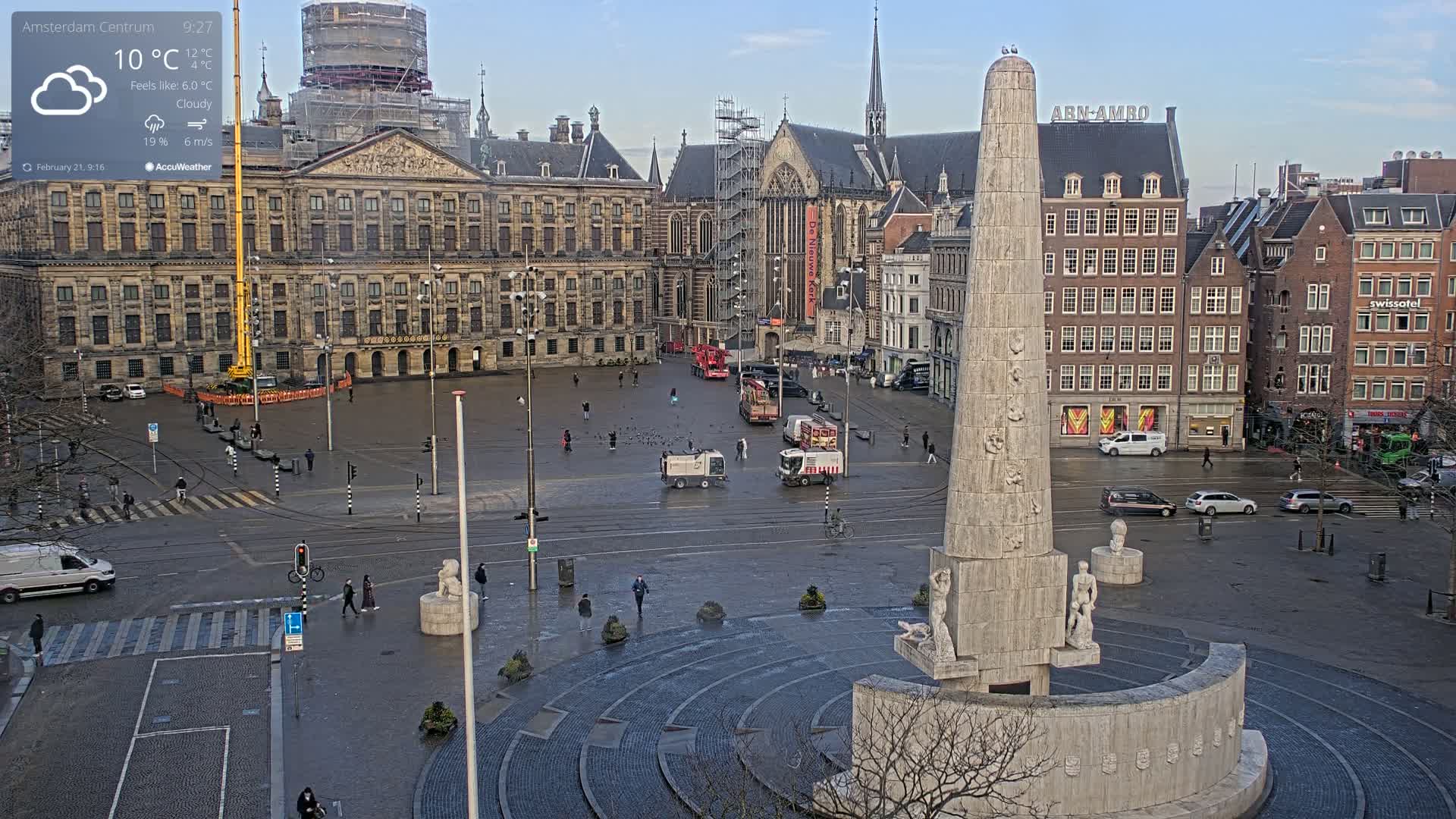 A cloudy day in a city square shows a large monument at the center, surrounded by buildings and a few pedestrians.