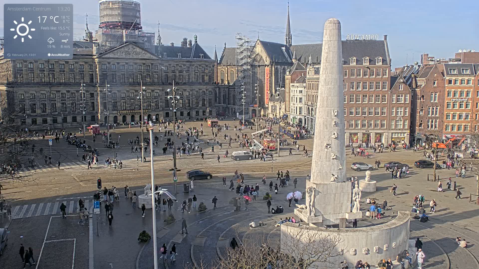 A sunny day in a large city square with many people, buildings, and a tall monument at its center.