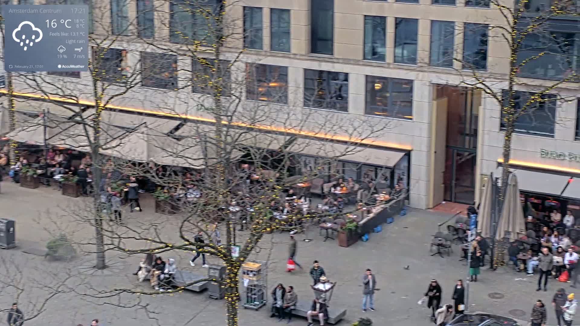 An overhead view of a city square with light rain, showing people gathered outside cafes under bare, string-light-adorned trees.