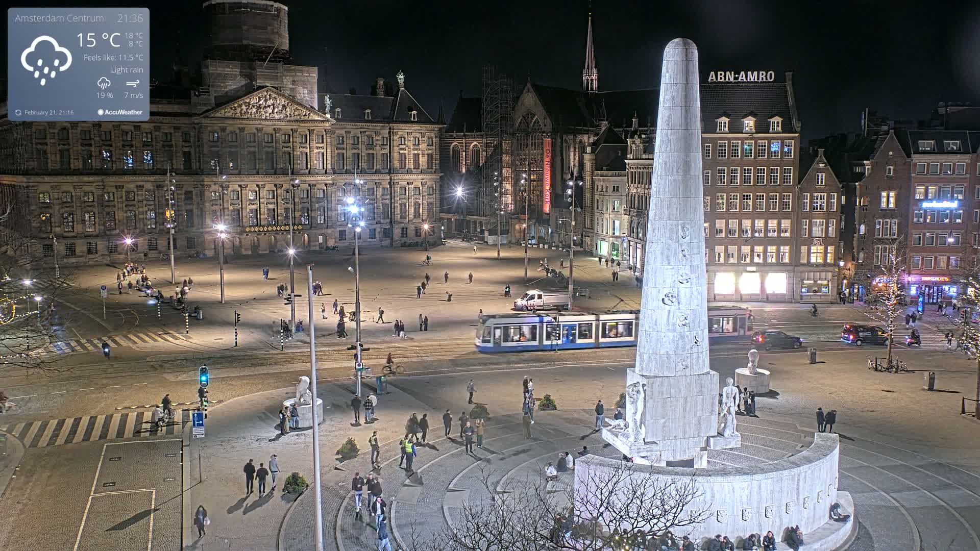 A nighttime, lightly raining view of a city square with a large monument in the center, surrounded by buildings and people, with a streetcar visible in the background.