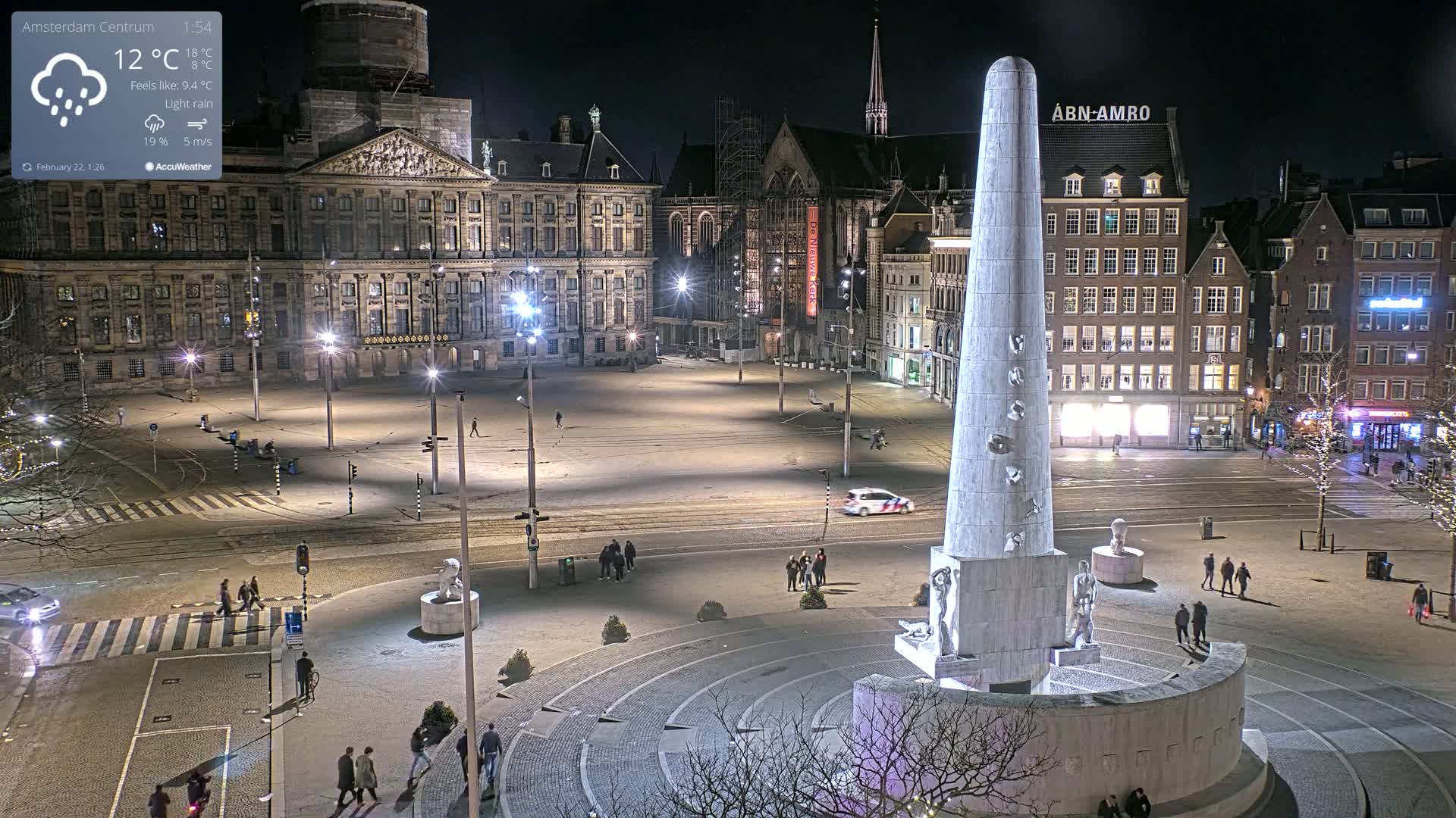A nighttime, lightly raining view of a large, empty city square with a tall monument in the center, surrounded by buildings, including a large palace.
