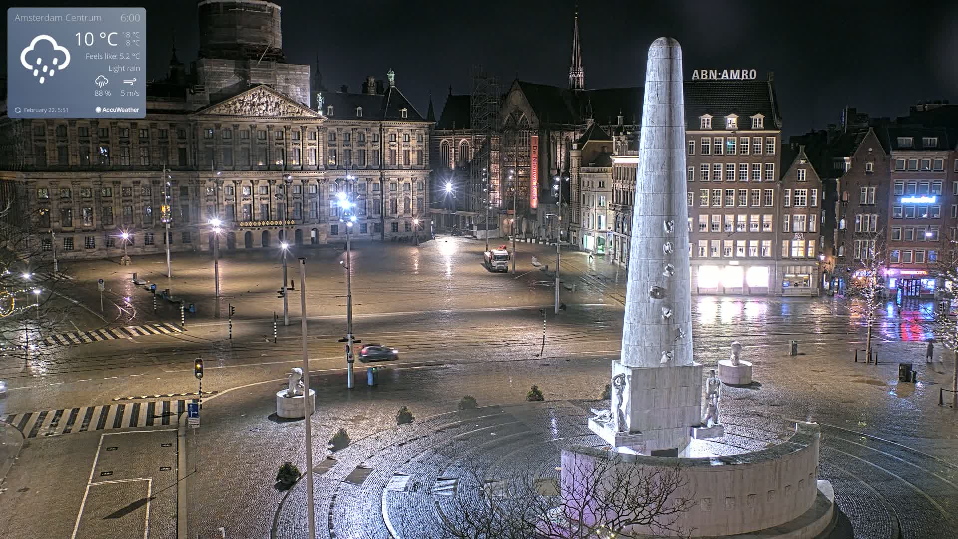 A nighttime, rain-slicked city square, centered on a tall, obelisk-like monument, with a palace and other buildings visible in the background under streetlights.