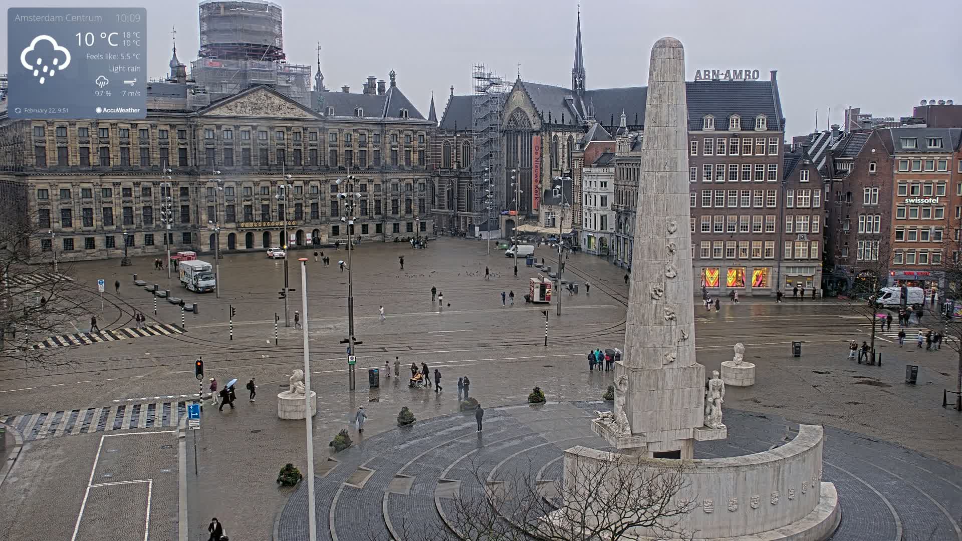 A large city square with a tall monument at its center is seen on a wet, overcast day with light rain.
