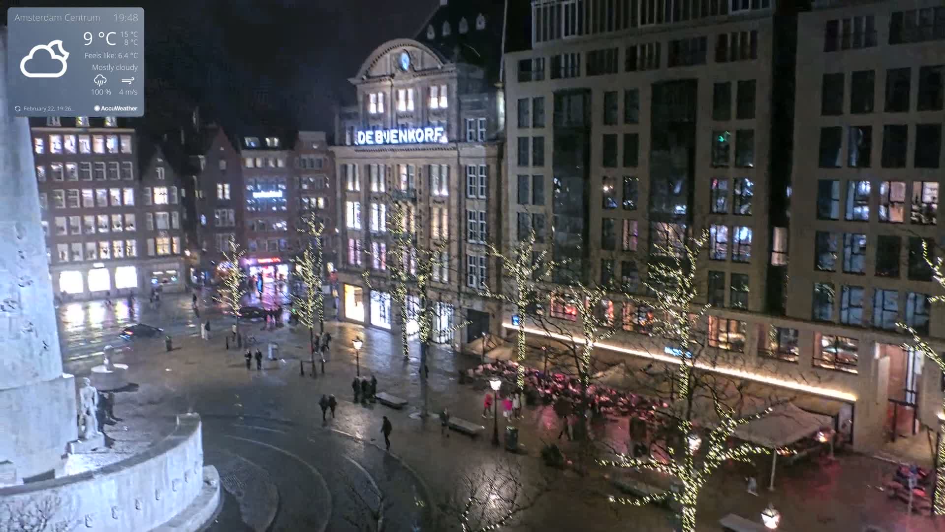 A nighttime, mostly cloudy view of a city square with illuminated buildings and trees decorated with lights, wet pavement, and sparse pedestrian traffic.