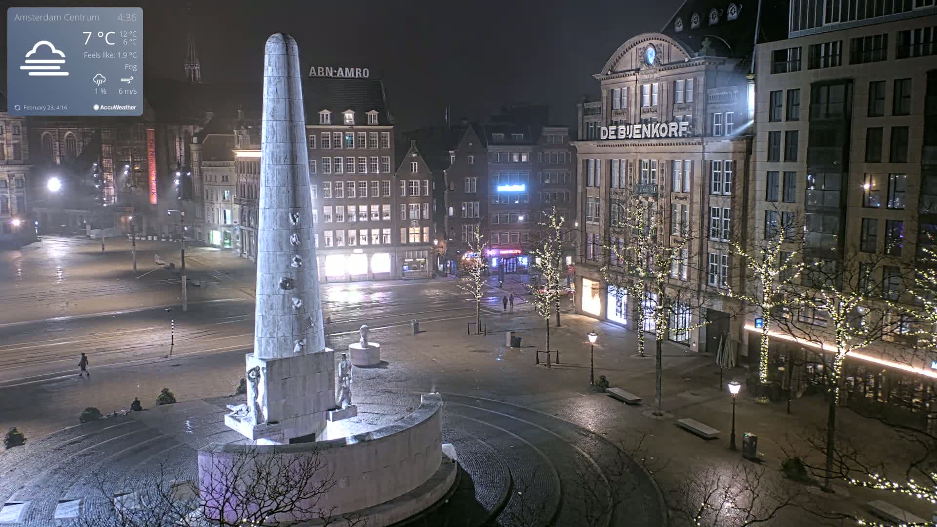 A nighttime, foggy city square with a tall monument at its center, surrounded by buildings illuminated with lights.