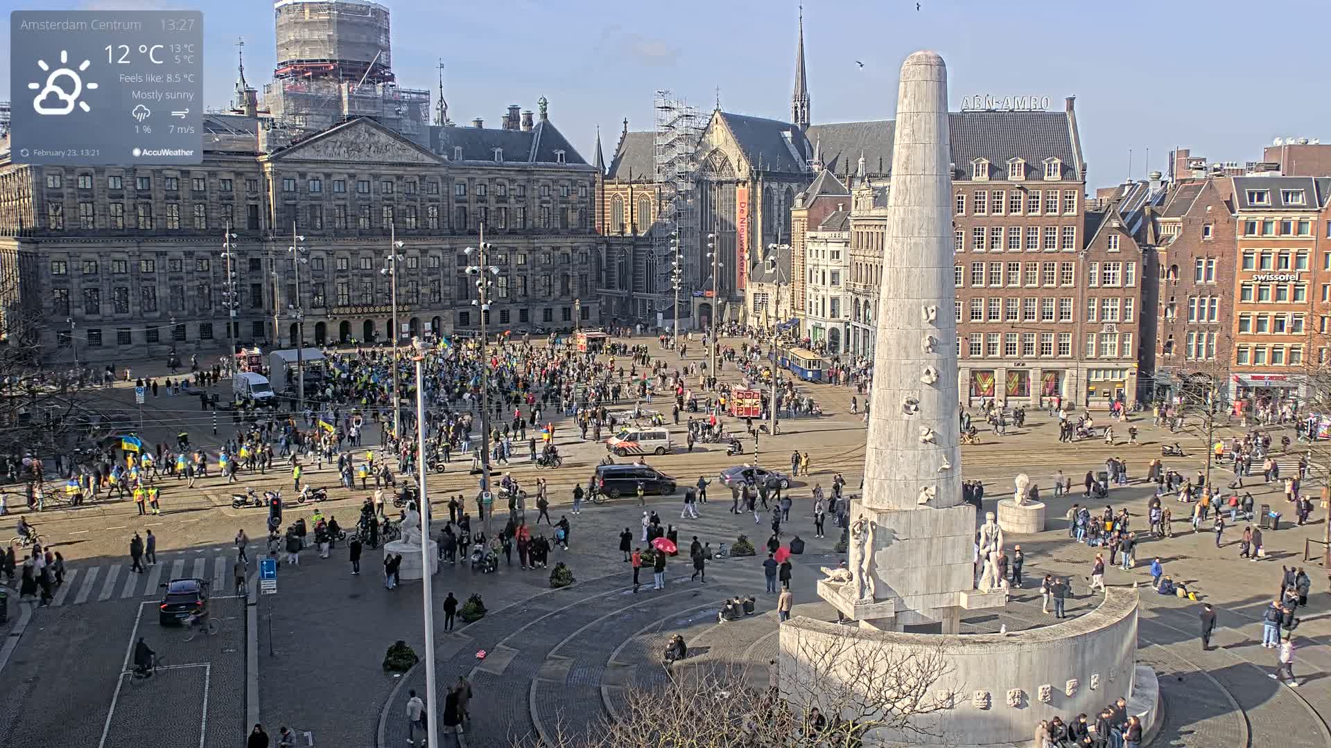 A mostly sunny day in a large city square, with many people gathered around a tall monument, and several buildings surrounding the square.