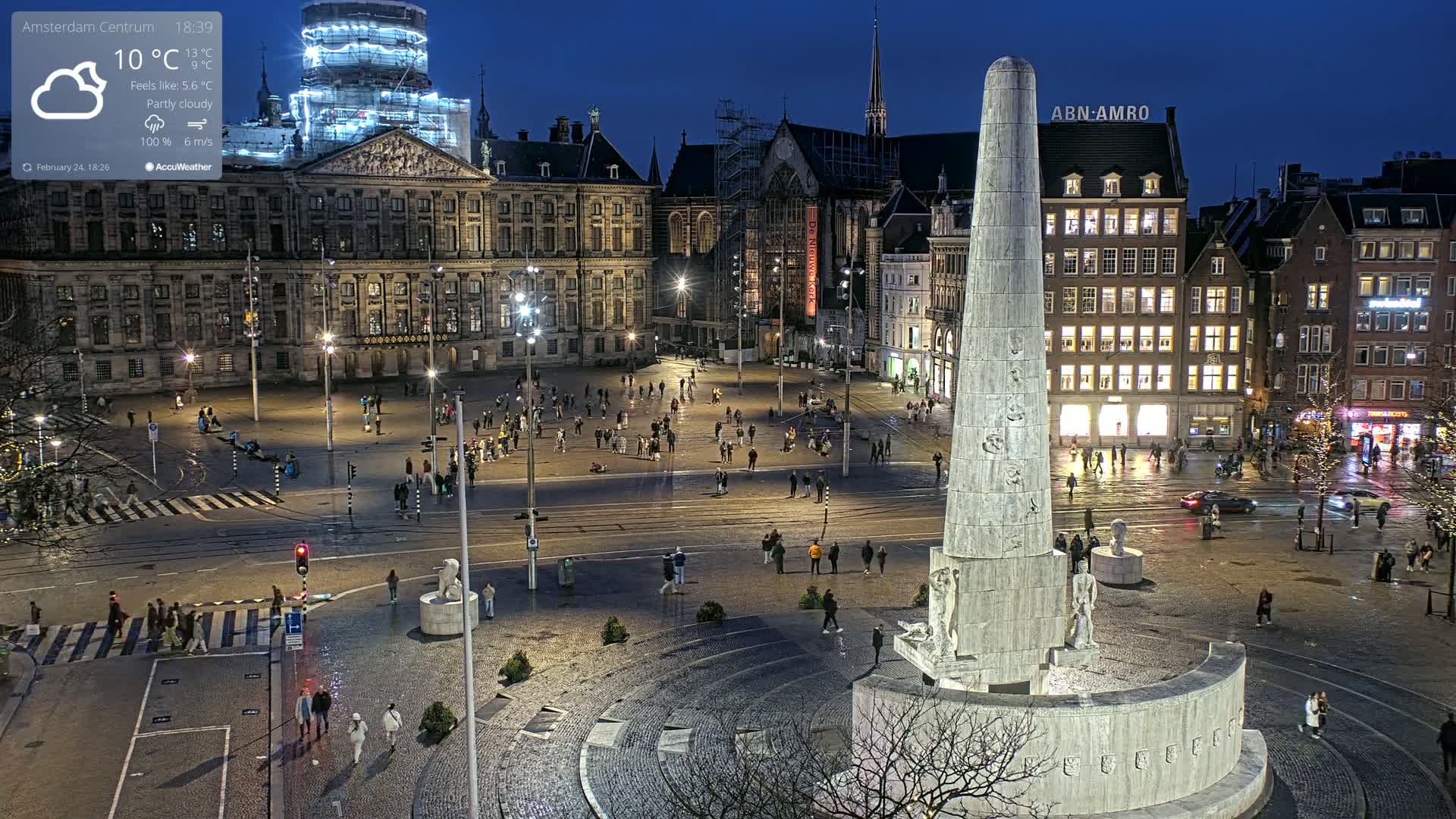 A nighttime, partly cloudy view of a large, illuminated city square with a tall monument at its center, surrounded by buildings and pedestrians.