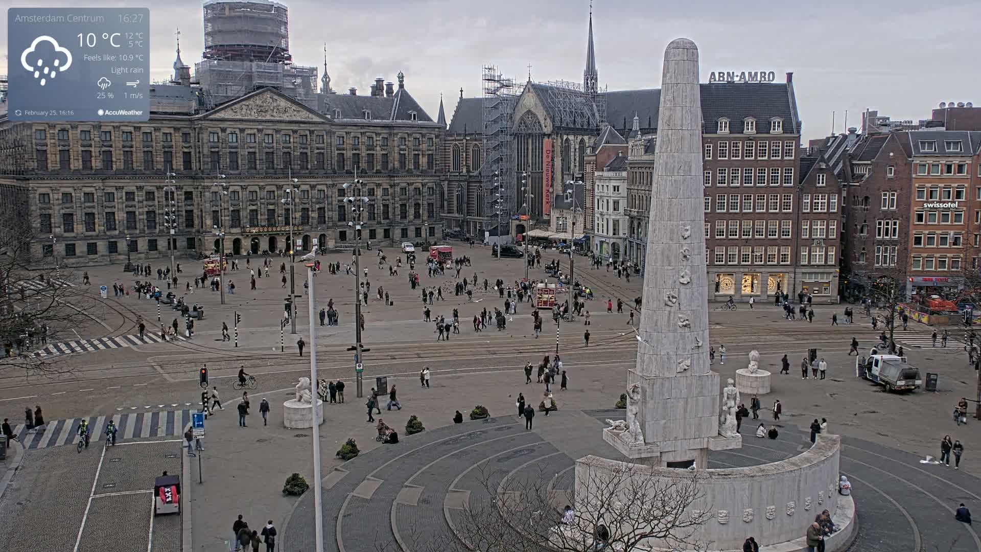 A large city square with a tall monument in the center, surrounded by historical buildings, under a light rain and overcast sky.
