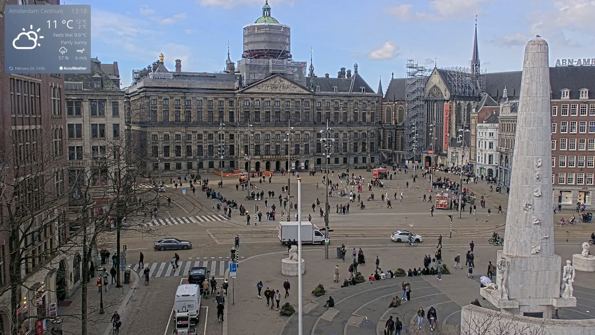 A partly sunny day in a large European city square shows a large palace undergoing renovations, several surrounding buildings, a monument, and numerous people walking around.