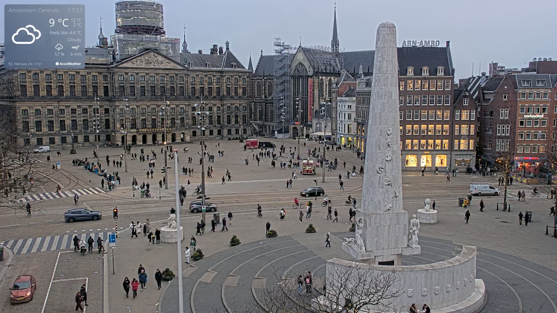 A cloudy, cool day in a large city square with a tall monument at the center, surrounded by historical buildings and people walking around.