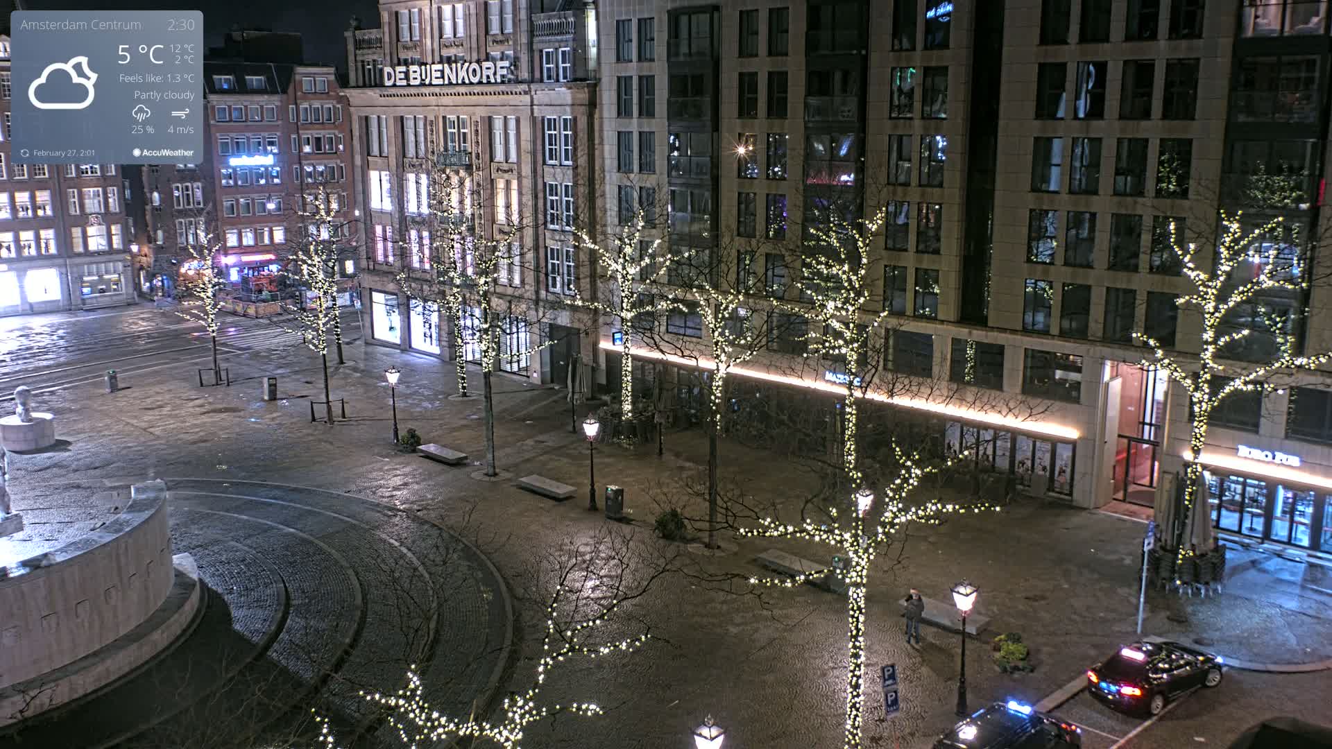 A nighttime, partly cloudy city square scene shows several buildings with illuminated windows, bare trees adorned with string lights, and a few cars parked on a wet cobblestone surface.