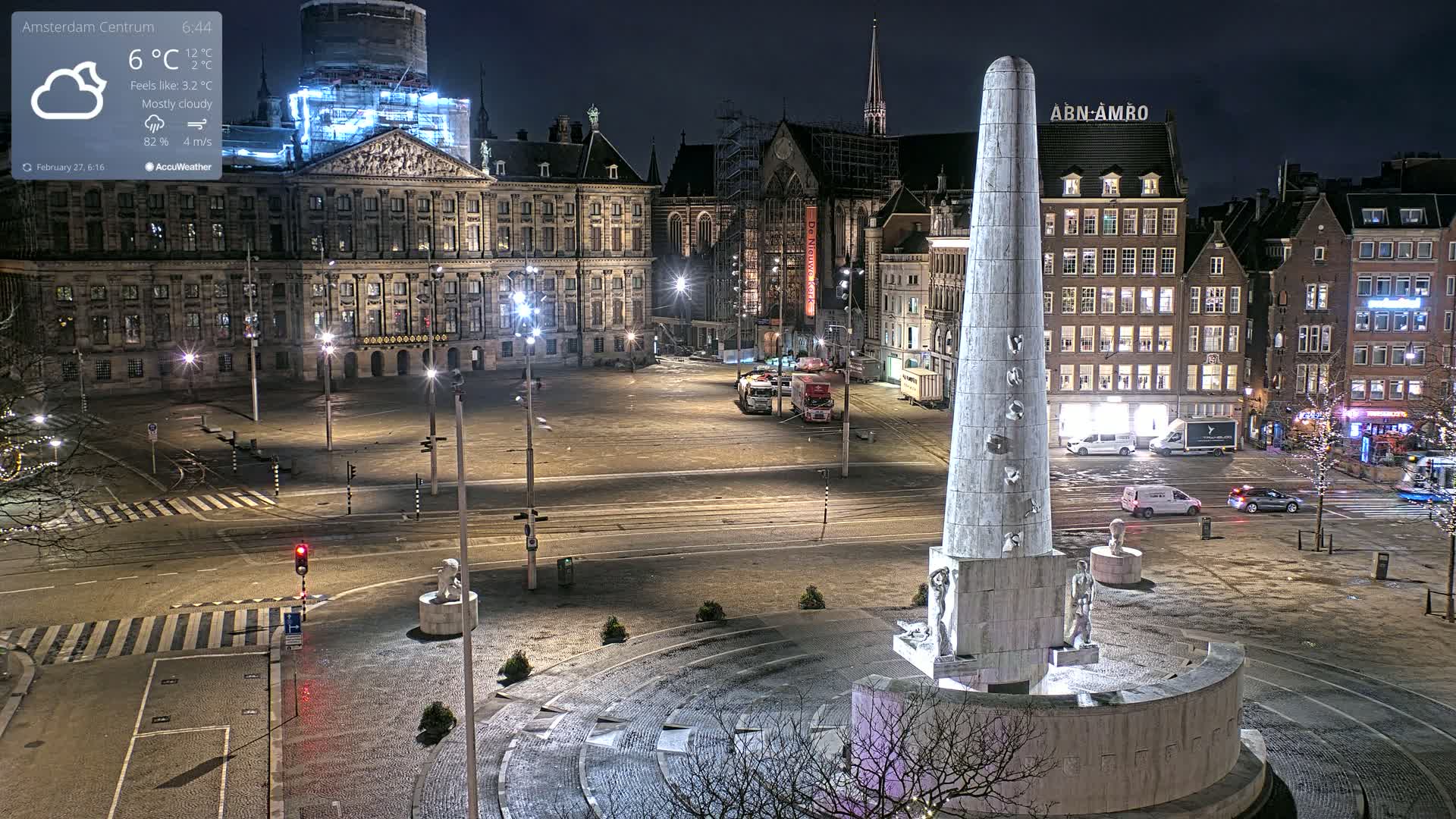 A mostly cloudy night scene shows a large, empty plaza with a tall monument in the center, surrounded by historical-looking buildings and illuminated streets.