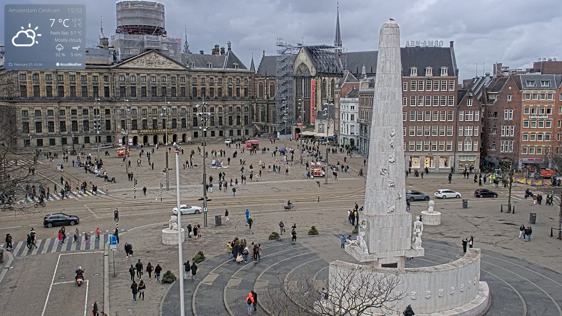 A mostly cloudy day reveals a large city square with a tall monument in the center, surrounded by historical buildings, people walking, and vehicles.