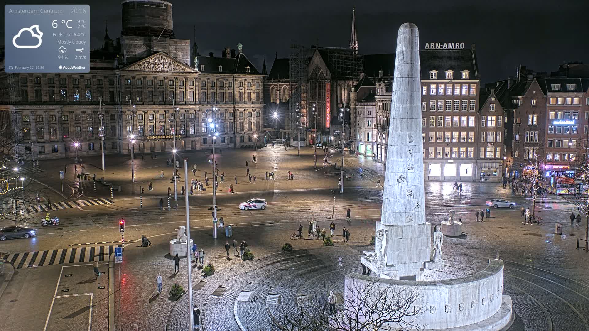 A nighttime, mostly cloudy view of a city square with a tall monument at its center, surrounded by historic buildings and sparsely populated with pedestrians.
