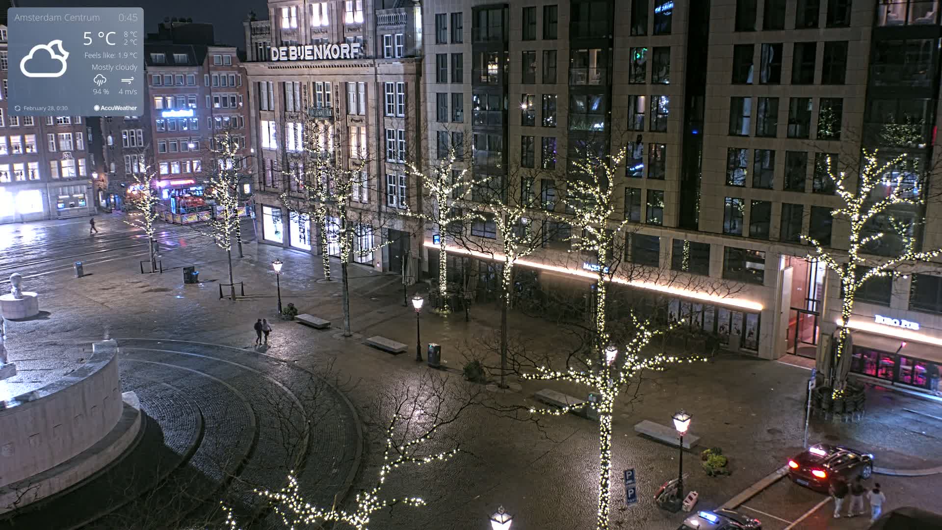A mostly cloudy night scene shows a city square with bare trees adorned with string lights, a fountain, and several buildings, one of which is brightly lit.