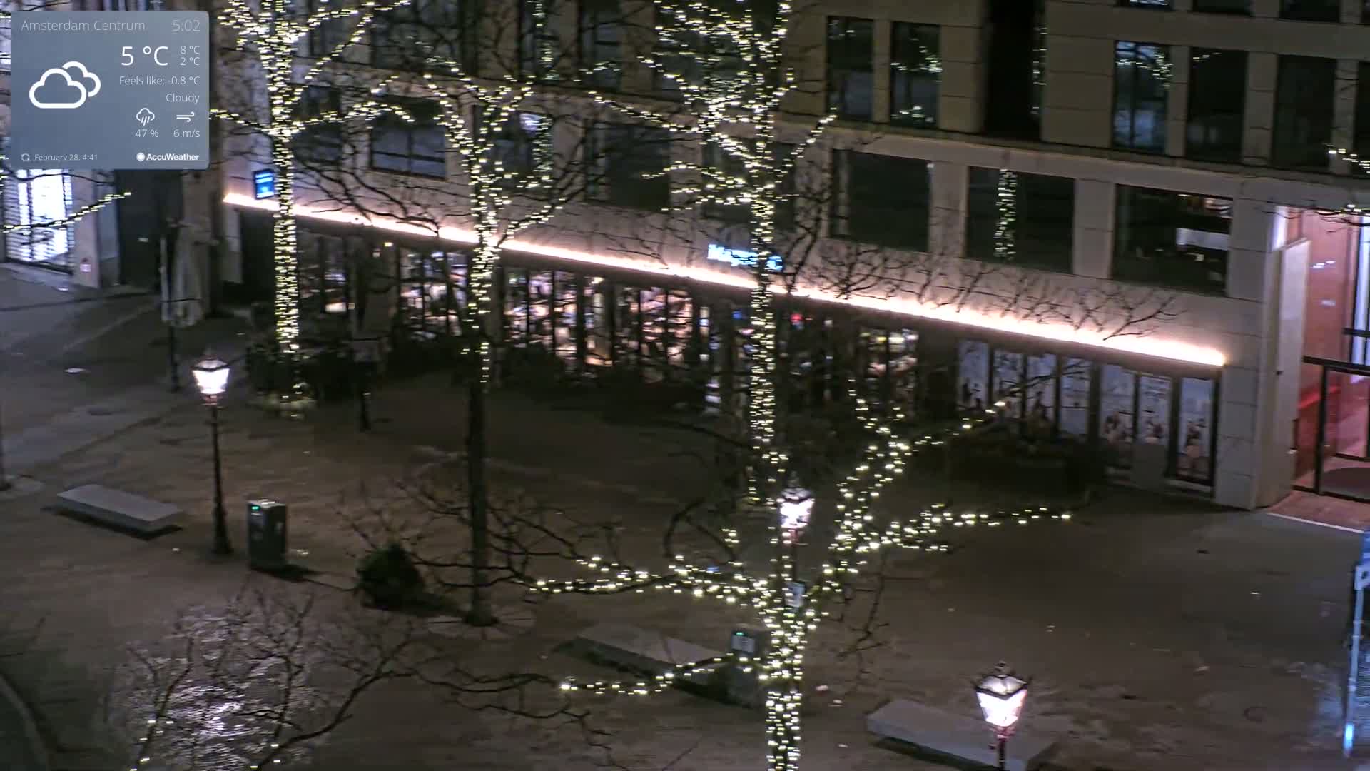 A nighttime view of a city square with bare trees decorated with string lights, a building with illuminated windows, and street lamps under cloudy skies.