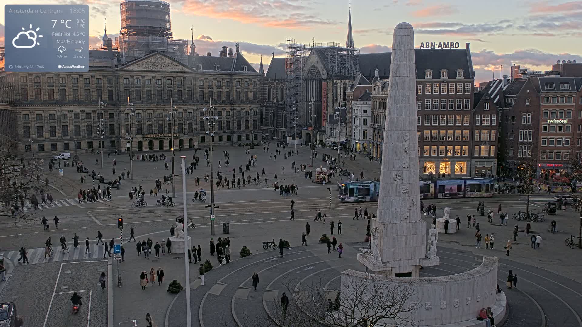 A mostly cloudy sunset illuminates a large city square with a monument at its center, surrounded by pedestrians and buildings, including a palace and a church under construction.