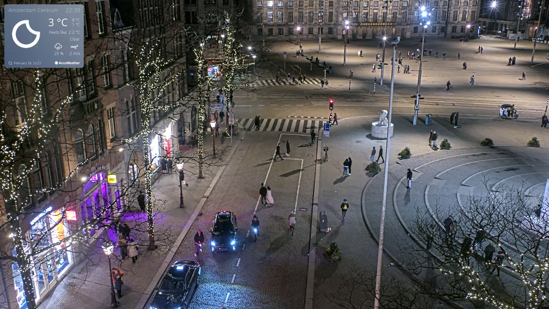 A nighttime, clear-weather aerial view of a city square with sparse pedestrian traffic, illuminated by streetlights and string lights on trees lining the streets.