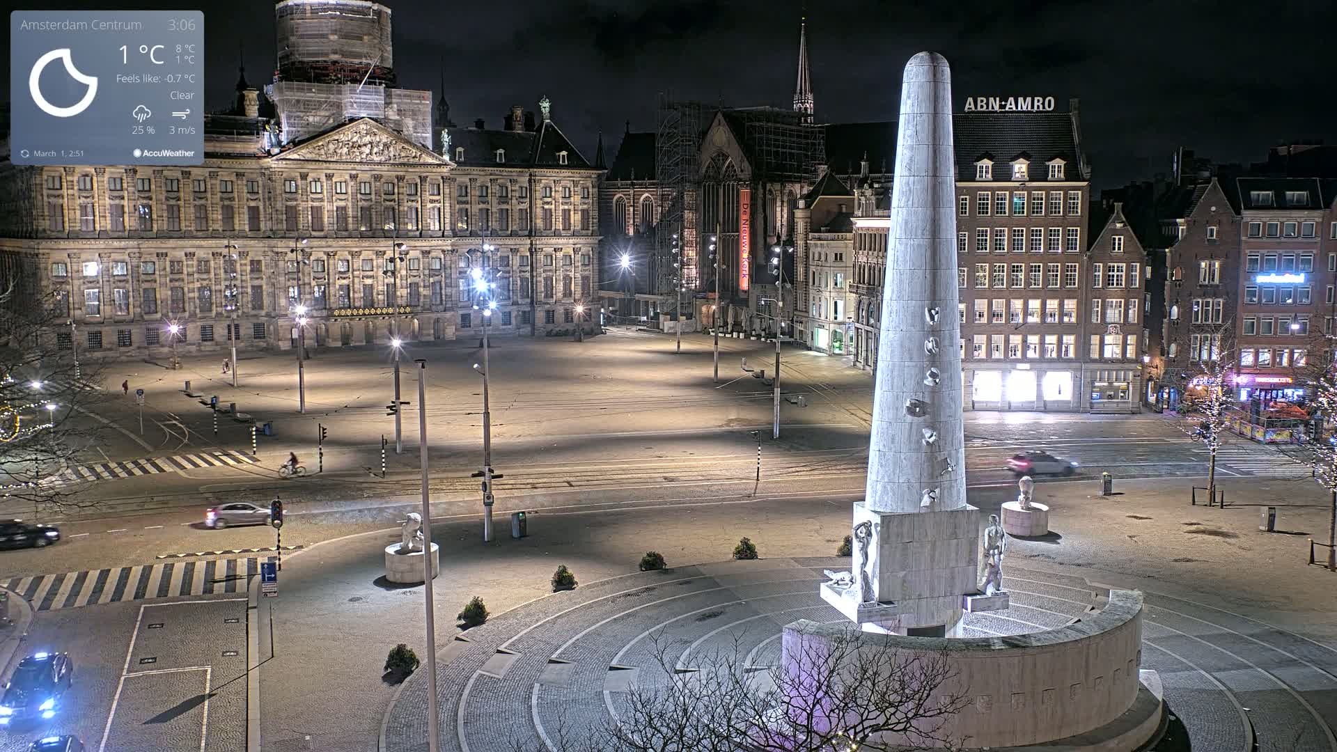 A nighttime, clear view of a large city square with a tall monument at its center, surrounded by historical buildings and sparsely populated streets.