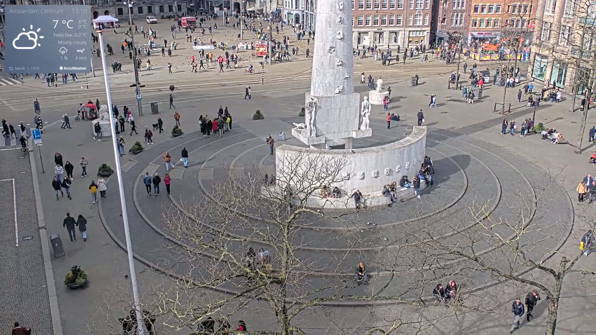 A mostly cloudy day shows many people gathered around a large, circular monument in a city square.