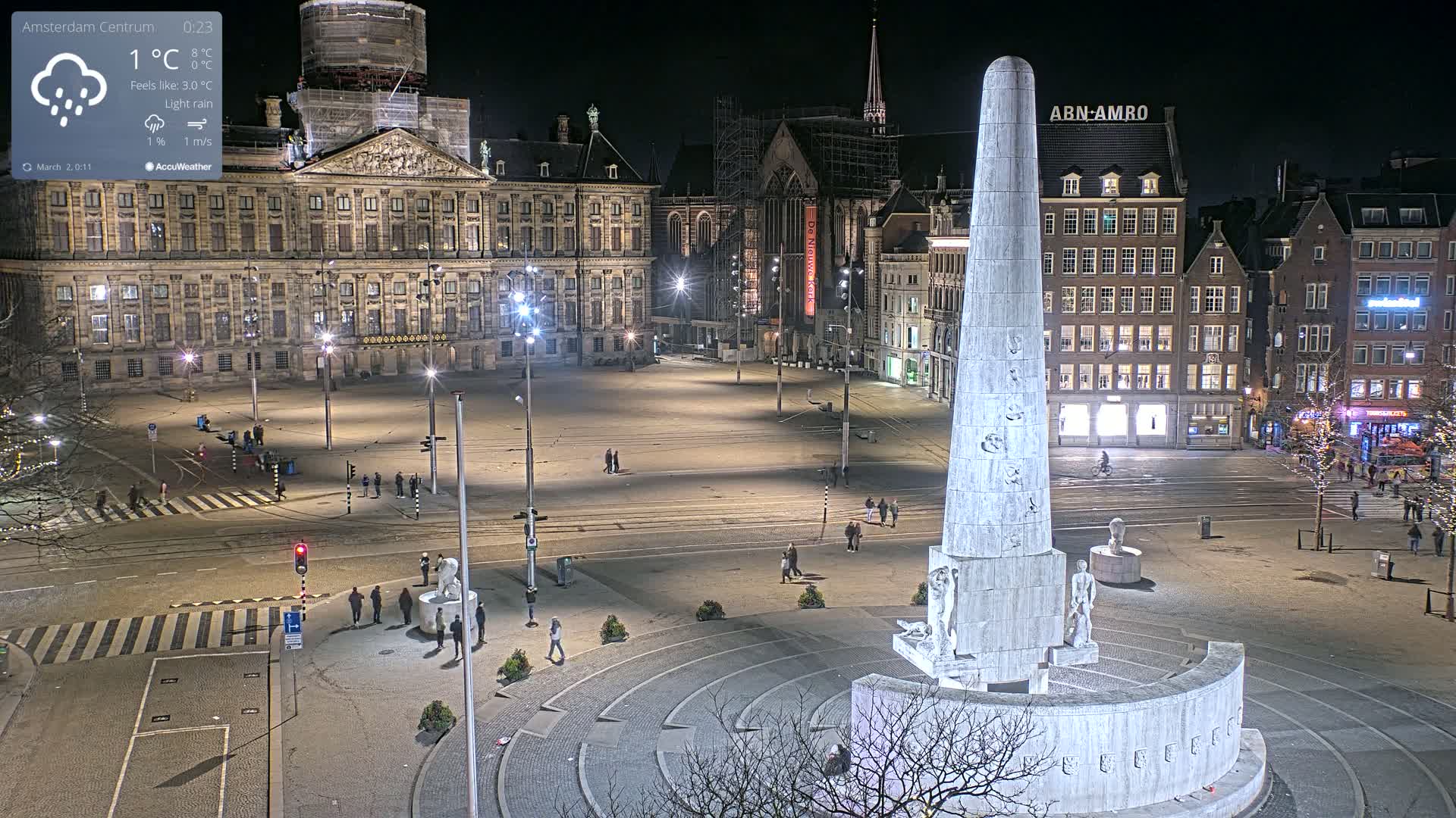 A nighttime view of a large, mostly empty city square with a tall monument at its center, under light rain.