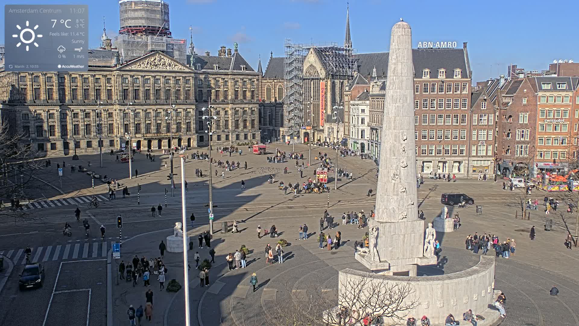 A sunny day in a large European city square, featuring a tall monument, historical buildings, and many pedestrians.