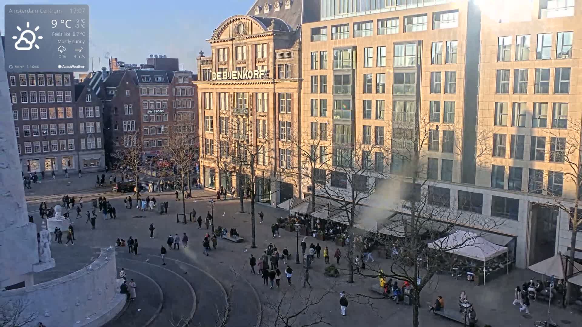 A mostly sunny city square, populated with pedestrians and outdoor seating areas, is surrounded by buildings under a partly cloudy sky.