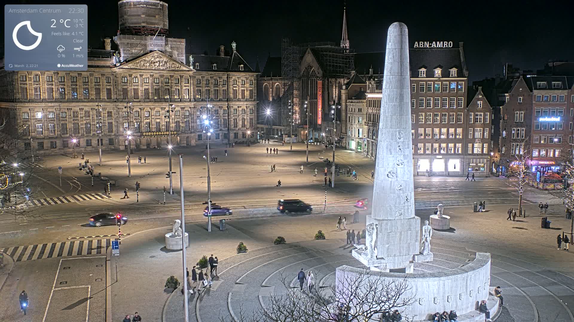A nighttime, clear view of a large city square with a tall monument at its center, surrounded by buildings and sparsely populated with people.