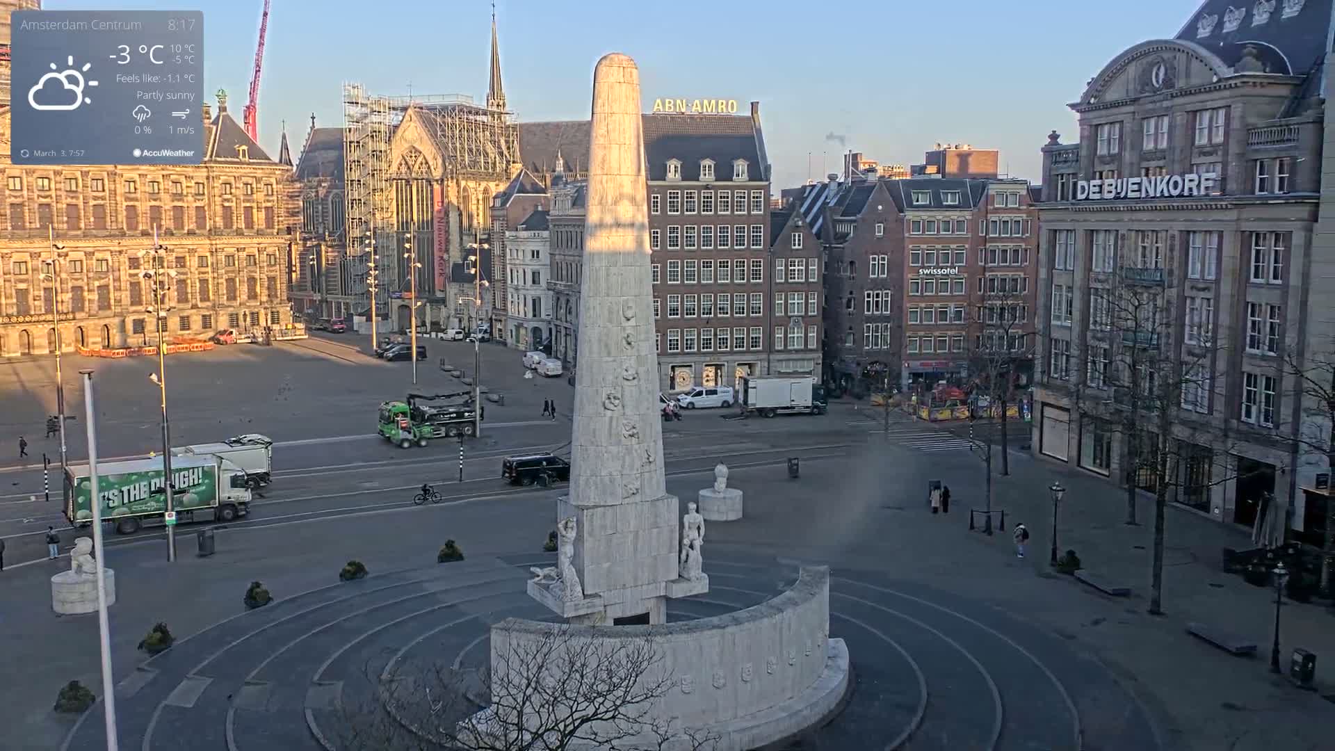 A partly sunny city square with a tall stone monument at its center, surrounded by several multi-story buildings.