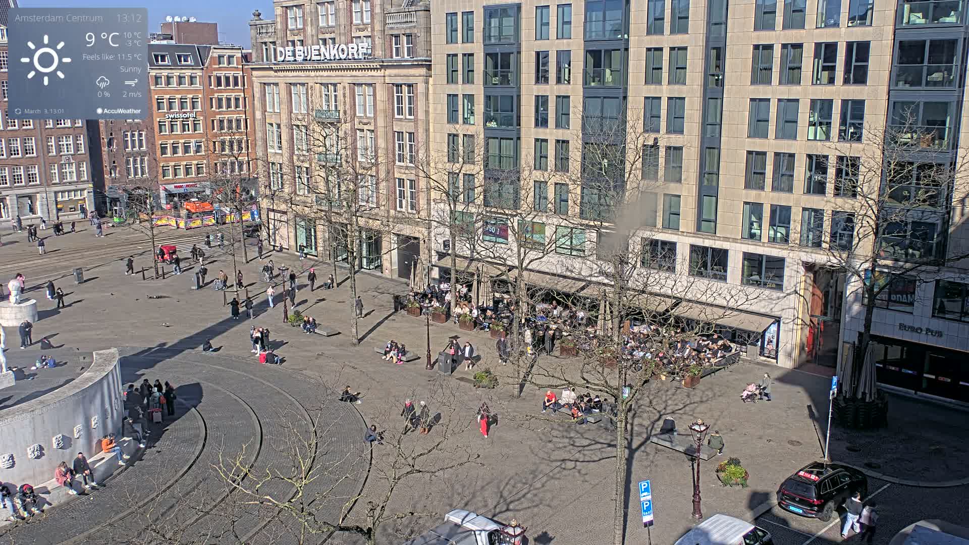 A sunny city square with many people sitting and walking around buildings, some of which have outdoor seating areas.