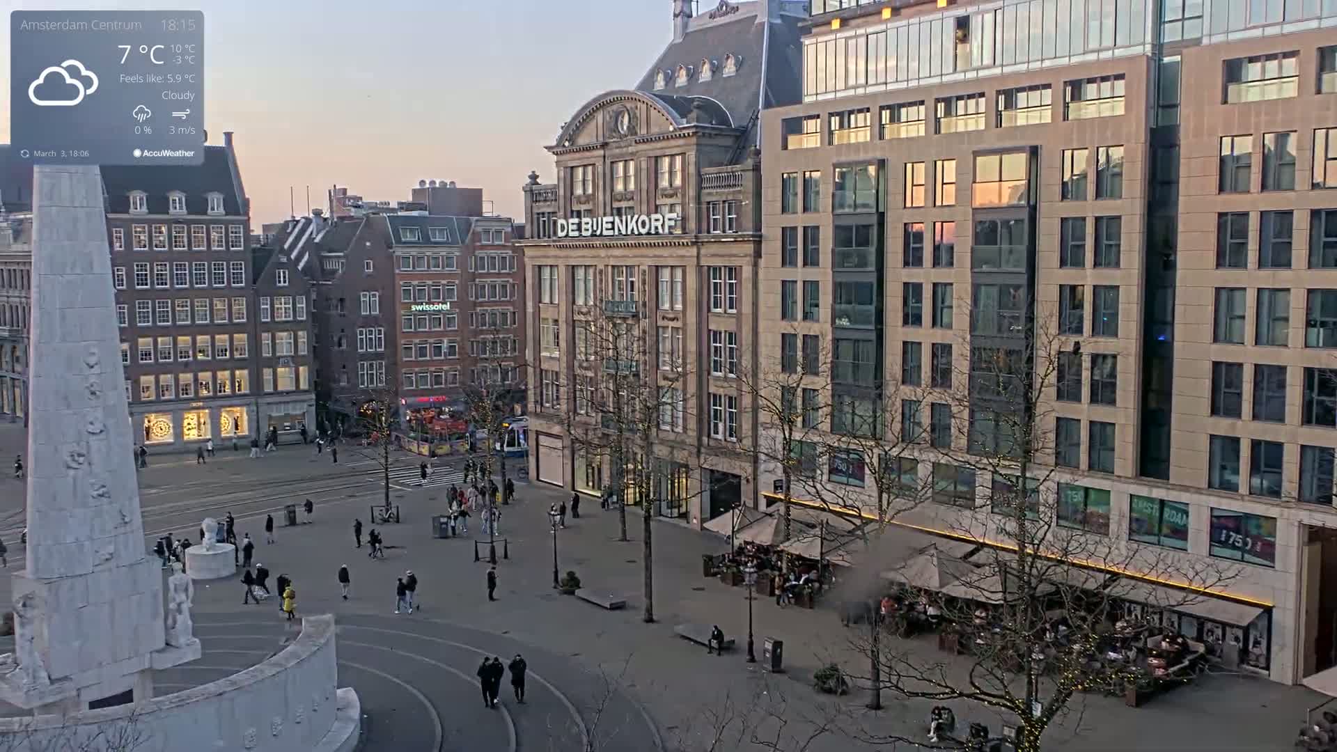 A cloudy, cool evening scene shows a city square with a monument, pedestrians, and buildings, including a large department store.
